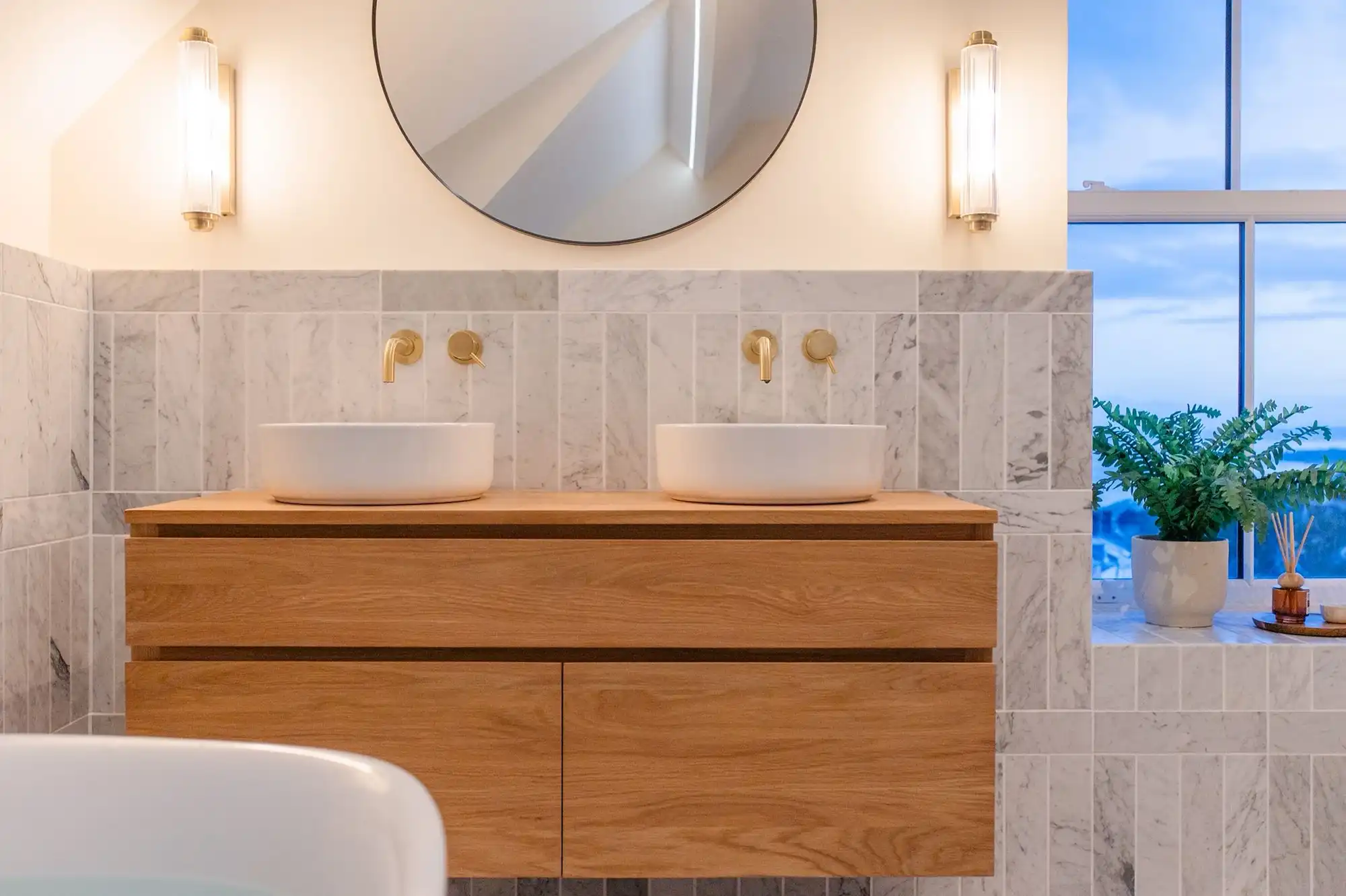 Modern bathroom with dual white vessel sinks on a wooden countertop, gold faucets, marble tiles, and a round mirror above.