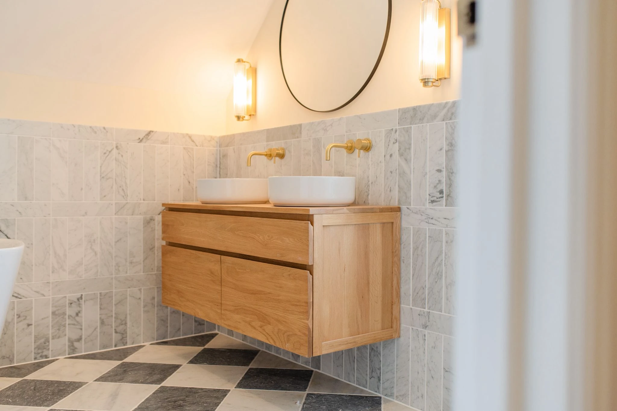 Stylish bathroom featuring a wooden vanity with two sinks, gold faucets, marble walls, and patterned floor tiles.