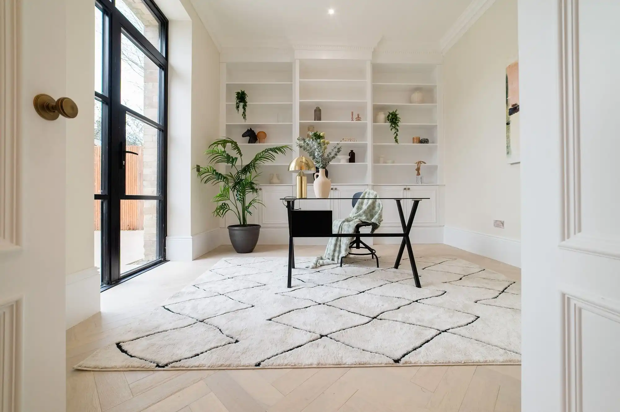 Modern home office featuring a black desk, cozy decor, and large windows with natural light. Decorated with plants and a stylish rug.