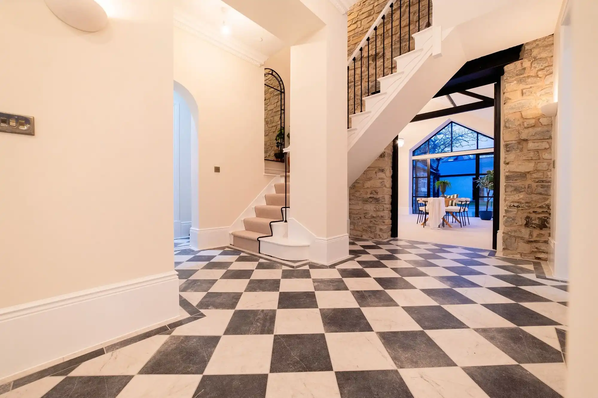 Spacious entryway featuring a checkered floor, a staircase, and stone walls, leading to a bright dining area with large windows.