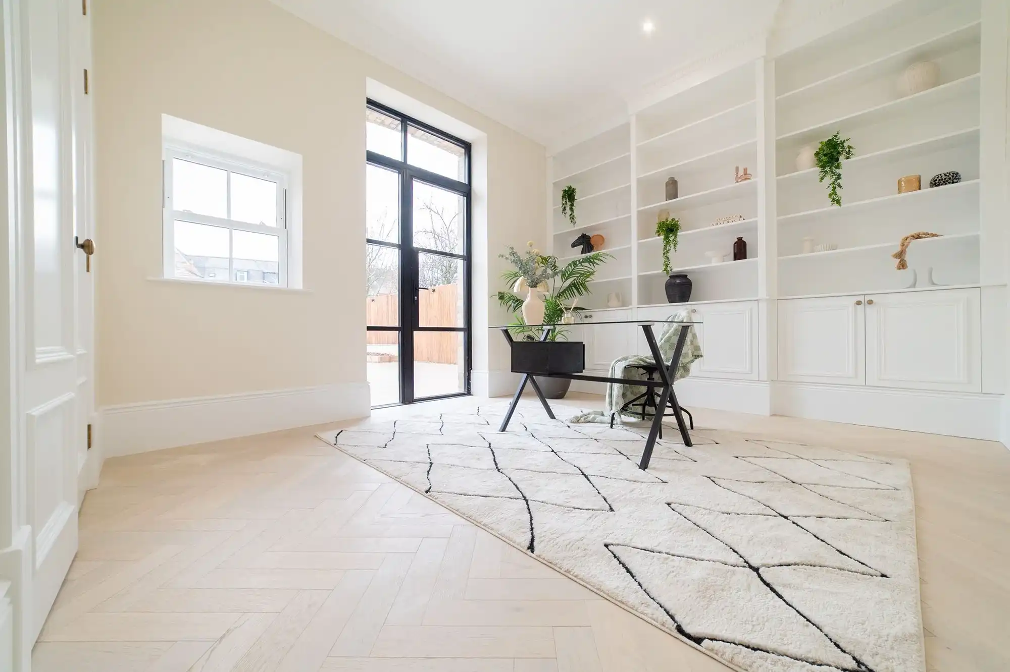 Bright, modern living space with a black glass door, white shelves, plants, and a patterned rug under a simple table and chair.